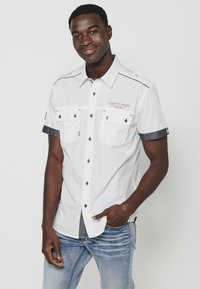Camisa blanca de manga corta con detalles en denim, dos bolsillos frontales y botones metálicos. Presenta un detalle del logo en el área del pecho.