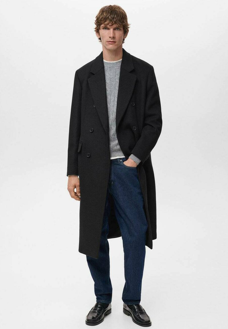 Hombre vistiendo un abrigo largo negro de doble botonadura, suéter gris, jeans azules y mocasines negros, de pie con una mano en el bolsillo frente a un fondo blanco.
