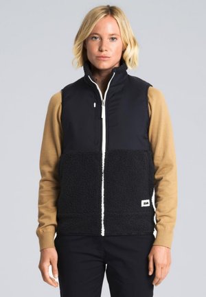Una mujer rubia luce un chaleco negro con cremallera delantera y la parte inferior de forro polar, sobre un jersey beige y ante un fondo gris liso.