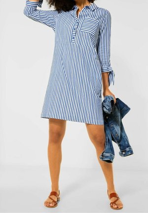 Robe chemise rayée en bleu et blanc. Présente un col, une poche avant et des manches roulées. Accompagnée de sandales à lanières marron et d'une veste en jean.