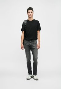 Μαύρη βαμβακερή μπλούζα, στενές γκρι τζιν παντελόνες και λευκά αθλητικά παπούτσια με μαύρες λεπτομέρειες. Το μοντέλο κρατάει μια τσάντα ώμου με λουράκι που έχει τρουκ.