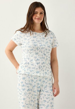 Nuori nainen, jolla on pitkät ruskeat hiukset, valkoinen lyhythihainen paita ja pienellä sinisellä kukkakuviolla varustetut housut, käsi lantiolla, hymyilee kevyesti.