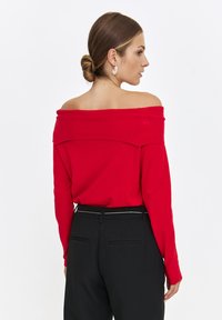 Roter schulterfreier Pullover mit geripptem Ausschnitt und langen Ärmeln, getragen über schwarzen, maßgeschneiderten Hosen mit schmalem Gürtel. Heller Farbkontrast.