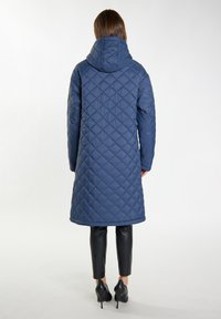 Cappotto lungo trapuntato navy con cappuccio, caratterizzato da un motivo a diamante e una consistenza liscia, abbinato a pantaloni neri aderenti e tacchi.