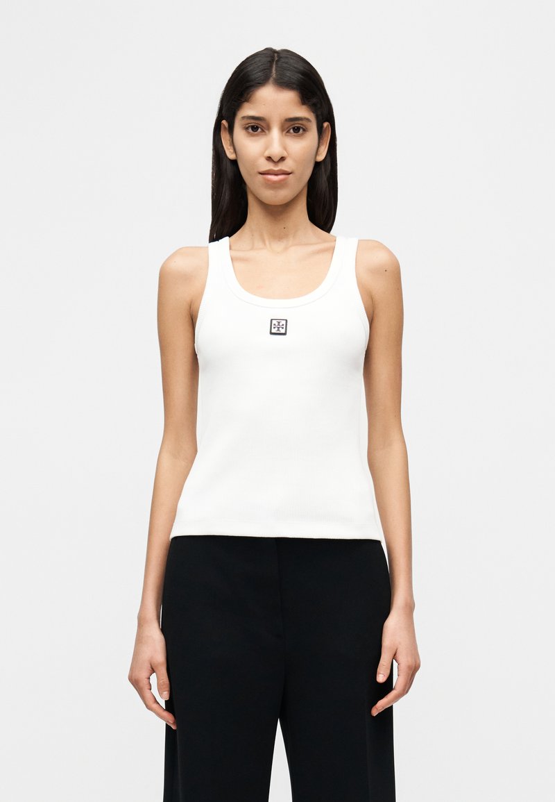 Λευκό ριμπ τιραντάκι με στρογγυλή  λαιμόκοψη, διακοσμημένο με ένα μικρό μαύρο λογότυπο στο κέντρο. Συνδυάζεται με μαύρο, φαρδύ παντελόνι.