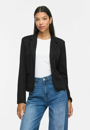 Femme aux longs cheveux foncés portant un blazer noir sur un top blanc et un jean bleu, debout devant un fond clair uni.