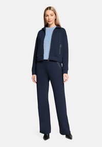 Navy blue zip-up sweatshirt with two side pockets and black accents, paired with a blue and white striped long-sleeve top and wide-leg pants.