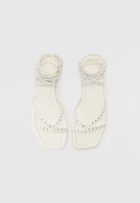 Sandales plates blanches avec plusieurs fines lanières ornées de petits clous en argent. Design à bout carré avec une texture lisse et mate.