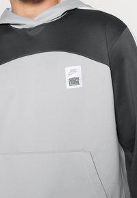 Šedá a černá mikina s kapucí, která má kulatý výstřih, kapsu a texturovanou tkaninu. Obsahuje bílou nášivku Nike s textem "FORCE".