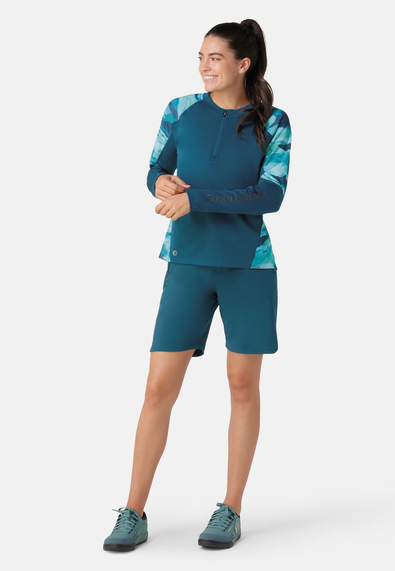 Teal aktivtøjssæt bestående af en langærmet top med lynlås i halsen og mønstrede ærmer; parret med matchende shorts og sneakers.