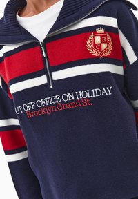 Granatowy i czerwony sweter z paskami, z prążkowanym kołnierzem, zamkiem błyskawicznym do połowy oraz haftowanymi detalami tekstowymi i logo. Miękka tekstura.