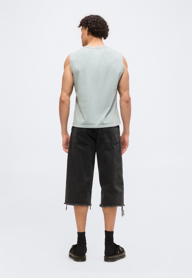 Hombre con pelo rizado que lleva una camiseta sin mangas gris claro, pantalones cortos negros deshilachados hasta la rodilla, calcetines negros y zapatos negros, de espaldas.
