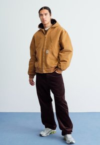 Bruine hooded jacket met een ritssluiting aan de voorkant, gecombineerd met donkerbruine corduroy broek. Model draagt lichte sneakers en staat op een blauwe ondergrond.