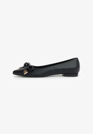 Scarpa piatta nera con punta appuntita in materiale strutturato, fiocco decorativo e dettagli in metallo dorato nella parte frontale, tacco basso, isolata su sfondo bianco.