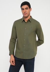 Olivzaļš garroku krekls ar pogājamu apkakli, priekšējām pogām un smalku logotipu augšējā kreisajā krūtīs. Gluda auduma tekstūra.