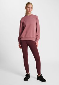 Lyserød sweatshirt med rund hals og lille logo på brystet, parret med ribbede bourgogne leggings og sorte sportsko.