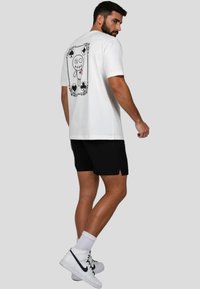 Camiseta blanca de algodón con un diseño gráfico en la parte posterior, que representa un personaje de dibujos animados enmarcado por símbolos de cartas de juego. Combinada con pantalones cortos negros.