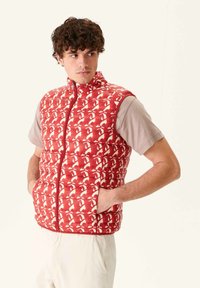 Gilet trapuntato rosso con motivo astratto bianco, colletto alto, zip frontale completa e tasche laterali, indossato sopra una camicia beige a maniche corte.