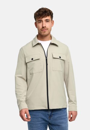 Ung mann med kort krøllete hår iført en beige jakke med glidelås og frontlommer med klaff, samt blå jeans, stående mot en ensfarget bakgrunn.