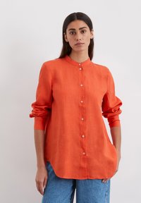 Oranžinė lininė marškinė su mandarinų apykakle, ilgomis rankovėmis, mygtukų uždarymu ir išlinkusia apačia. Dėvimi su mėlynomis džinsinėmis kelnėmis.