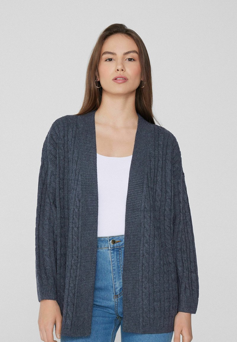 Dunkelgrauer, gestrickter Cardigan mit einem Zopfmuster, offener Vorderseite und langen Ärmeln. Getragen über einem weißen Oberteil und kombiniert mit blauen Jeans.