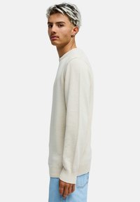 Vaaleanbeige poolopusero, jossa resorit hihansuissa ja helmassa, ja jossa on teksturoitu neulekuvio. Yhdistetty vaaleansinisiin farkkuihin.