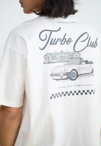 Balta kokvilnas T-krekls ar grafisku attēlu, kurā redzams kabriolets un ēka, ar tekstiem "Turbo Club" un "911 Turbo" pelēkā krāsā.