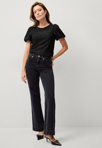 Schwarzes Spitzenoberteil mit Puffärmeln, kombiniert mit hoch taillierten, schwarzen Schlagjeans. Das Model trägt schwarze spitze Absätze und silberne Ohrringe.