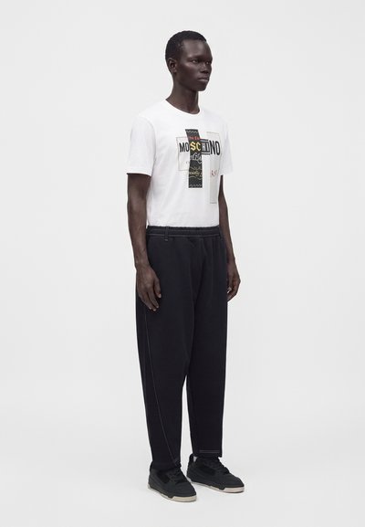 Άνδρας που φοράει λευκό μπλουζάκι Moschino με γραφικό σχέδιο, φαρδιά μαύρη παντελόνα και μαύρα αθλητικά παπούτσια με φωτεινές σόλες σε ένα απλό λευκό φόντο.