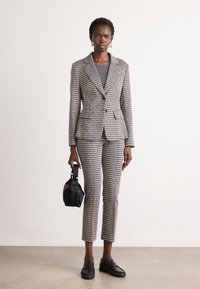 Gray and black patterned suit with a fitted blazer and cropped pants. Model holding a small black handbag, wearing flat black shoes.