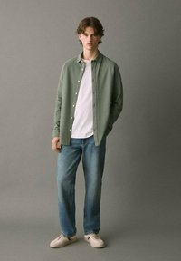 Camicia button-up verde sopra una t-shirt bianca, abbinata a jeans azzurri chiari e scarpe beige. Sfondo semplice.