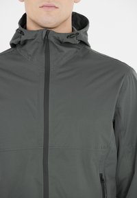 Donkergrijze lichtgewicht jas met een hoge kraag en een verstelbare capuchon. Voorzien van een voorrits en zijzakken. Slanke, gladde textuur.