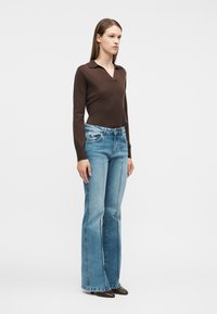 Γυναίκα όρθια που φορά μακρυμάνικη μπλούζα σκούρου καφέ χρώματος και ανοιχτό μπλε τζιν ψηλόμεσο με φαρδιά μπατζάκια και ραφές, καθώς και καφέ μυτερά παπούτσια.