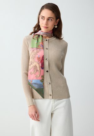 Vrouw draagt een beige geribbelde knoopcardigan, witte broek en een kleurrijke sjaal met patroon om haar nek geknoopt, kijkt opzij.