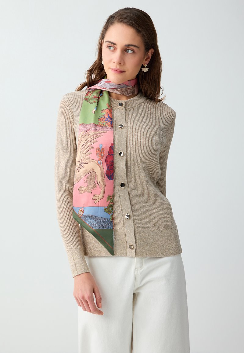 Femme portant un cardigan beige côtelé à boutons, un pantalon blanc et une écharpe colorée à motifs nouée autour du cou, regardant sur le côté.