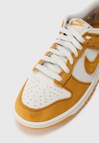 Žlté a biele tenisky z semišu a sieťoviny, s perforovanou špičkou, bielymi šnúrkami a výrazným oranžovým logom Nike swoosh.