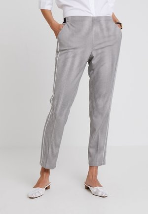 Kobieta ubrana w białe i czarne wąskie spodnie w kratę z białymi bocznymi paskami, białą koszulę zapinaną na guziki oraz białe plecione buty wsuwane.