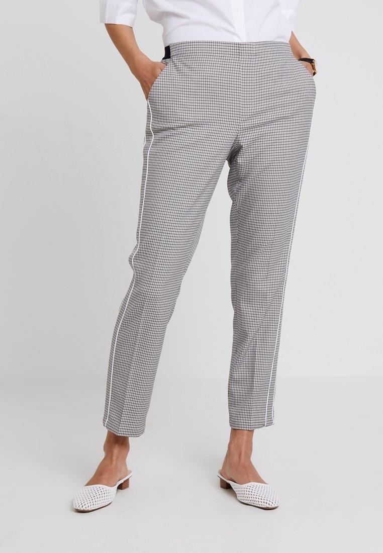Femme portant un pantalon slim à carreaux blancs et noirs avec des bandes latérales blanches, une chemise blanche boutonnée et des chaussures blanches tissées à enfiler.