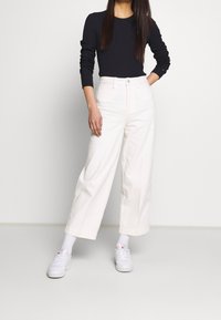 Valkoiset leveälahkeiset farkut, joissa on korkea vyötärö ja sivutaskut, yhdistetty mustaan pitkähihaiseen toppiin ja valkoisiin tennareihin.