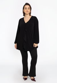 Blouse noire à manches longues avec décolleté en V, dotée d'un design front ruffle, associée à des leggings noirs évasés et des escarpins bordeaux à bouts pointus.