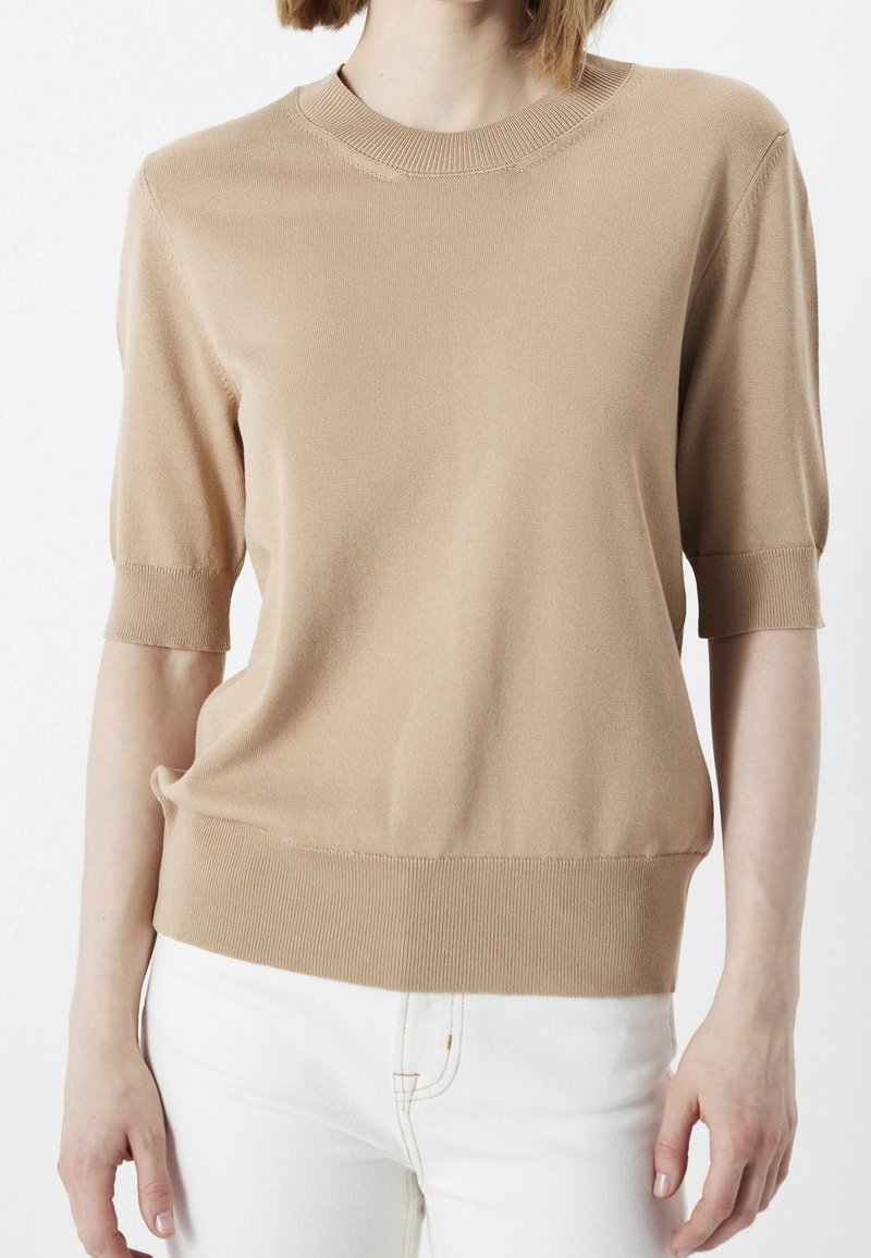 Femme portant un pull en maille beige à manches courtes associé à un pantalon blanc, montrée des épaules aux hanches sur un fond uni.