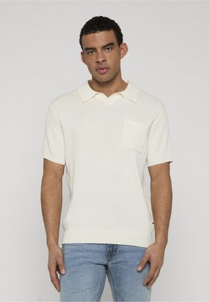 Muž nosící krémově bílou polokošili s krátkými rukávy a žebrovaným vzorem, s náprsní kapsou, v kombinaci se světle modrými džínami, stojící před jednoduchým pozadím.