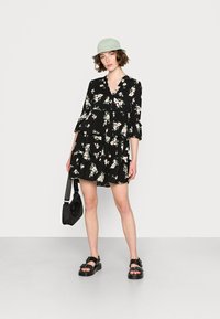 Vestido preto com padrão floral, mangas onduladas e design em camadas. A modelo usa um chapéu estilo bucket verde claro e sandálias pretas. Inclui uma bolsa preta sólida.