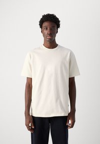 Tricou din bumbac light beige, cu mâneci scurte, gât rotund și croială lejeră. Prezintă un design minimalist, fără logo-uri sau modele vizibile.