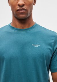 Tricou turquoise cu guler rotund, realizat din bumbac, având un mic logo alb "Marc O'Polo DENIM" în zona pieptului stâng.