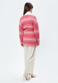 Overmaatse roze gradient trui met geribbelde textuur, met een ceintuur in de taille en lange mouwen, gecombineerd met crèmekleurige wijde broek en beige hakken.