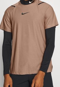 Camiseta atlética de manga corta, color marrón claro, con un logo negro de Nike, presenta un escote redondo y mangas raglán sobre una capa base de manga larga negra.