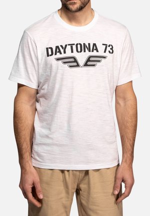 Homme portant un T-shirt blanc avec un texte noir "DAYTONA 73" et un emblème ailé, associé à un pantalon beige, debout devant un fond uni.