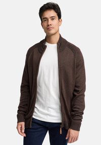 Brun strikket cardigan med lynlåslukning foran, ribbet krave og manchetter samt en diskret struktur. Båret over en hvid T-shirt og mørke jeans.