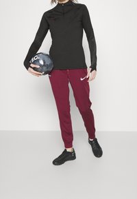 Schwarzes, langärmliges Sportoberteil mit Reißverschlusskragen, strukturierte Ärmel; kastanienbraune Jogginghose mit weißem Nike-Logo; schwarze Sportschuhe; hält einen schwarzen Ball.